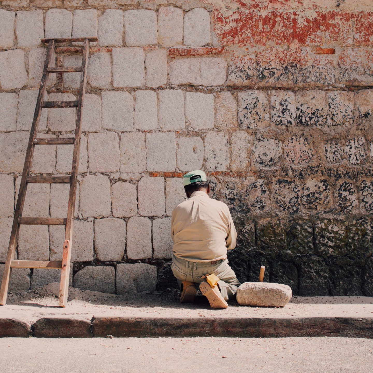 Main kneels in the sun against a wall and paints it white. A ladder leans to the side.